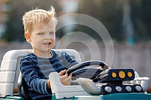 Cute little boy driving big electric toy car with steering wheel and having fun outdoors