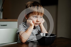 Cute little boy eating oatmeals for beakfast dreamily looking at the window. image with selective focus