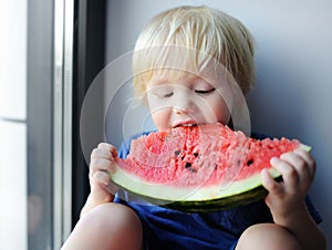 Cute little boy eating watermelon sitting on windowsill