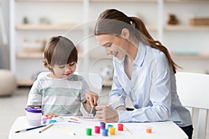 Cute little boy enjoying art classes, drawing picture with colorful paints, sitting at desk with teacher and laughing