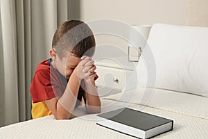 Cute little boy with hands clasped together saying bedtime prayer over Bible at home. Space for text