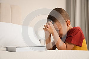 Cute little boy with hands clasped together saying bedtime prayer over Bible at home. Space for text