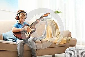 Cute little boy with headphones playing guitar on sofa in room