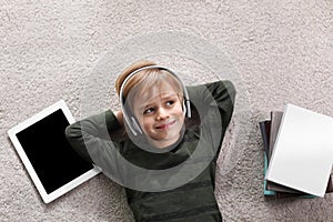 Cute little boy with headphones and tablet listening to audiobook on floor, flat lay