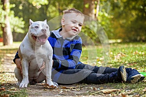 Cute little boy with his friend, five months old puppy of American Bully dog of white color, sitting in autumn park and smiling.