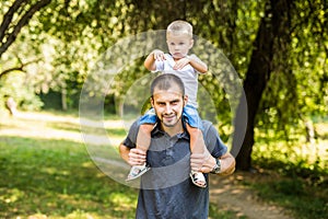Cute little boy sitting pickaback of his handsome young dad are looking at each other and smiling while resting in the park photo