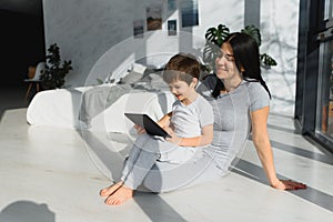 Cute little boy and his young mother using a digital tablet and smiling on the floor at home. Happy family with gadget