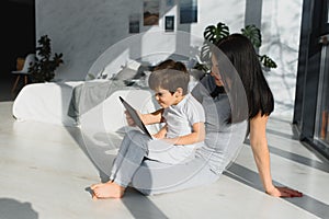 Cute little boy and his young mother using a digital tablet and smiling on the floor at home. Happy family with gadget