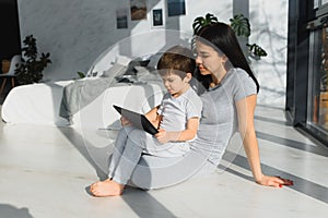 Cute little boy and his young mother using a digital tablet and smiling on the floor at home. Happy family with gadget