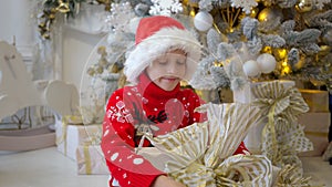 cute little boy holding big Christmas gift shaking it smiling on the background of elegant Christmas tree at home 4K