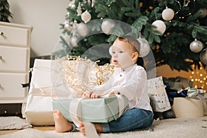 Cute little boy at home by the big Christmas tree looking for presents. Happy kid spend time on Holiday and enjoying