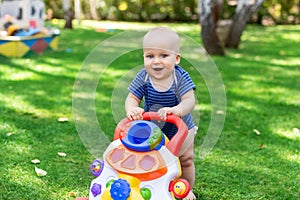 Cute little boy learning to walk with walker toy on green grass lawn at backyard. Baby laughing and having fun making