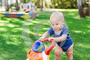 Cute little boy learning to walk with walker toy on green grass lawn at backyard. Baby laughing and having fun making