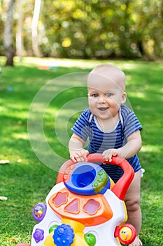 Cute little boy learning to walk with walker toy on green grass lawn at backyard. Baby laughing and having fun making first step a