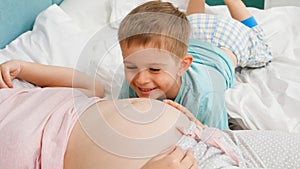 Cute little boy in pajamas listening to unborn baby in pregnant mother belly lying on bed. Concept of loving children