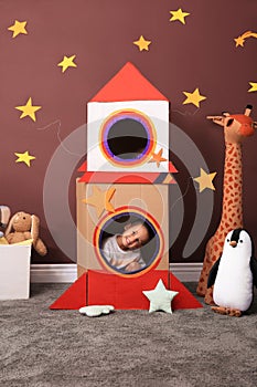 Cute little boy playing with cardboard rocket and toys at home. Child`s room interior