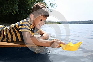 Cute little boy playing with paper boat on wooden pier near river