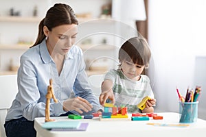 Cute little boy preschooler playing developing wooden games with professional woman psychologist at kindergarten