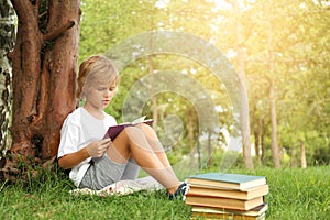 Cute little boy reading book on green grass near tree in park