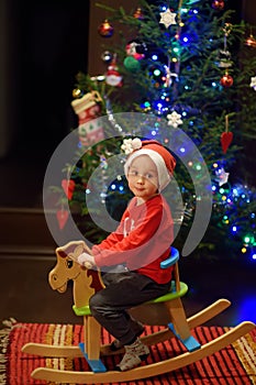 Cute little boy on rocking horse chair for children near decorated fir tree