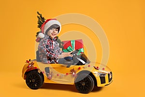 Cute little boy in Santa hat with Christmas tree and gift box driving children`s electric toy car on yellow background