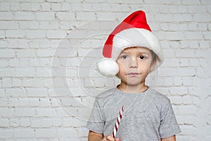 Cute little boy in Santa hat eating Christmas candy cane sweets on white bricks wall background