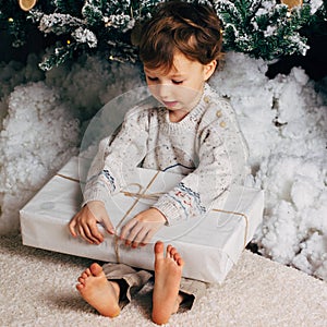 Cute little boy sitting with a box of gifts around the Christmas tree. Adorable blonde caucasian toddler. Square photo