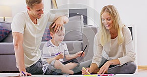 Cute little boy sitting on floor engaged in family activity paint in album together with young parents, happy loving mom