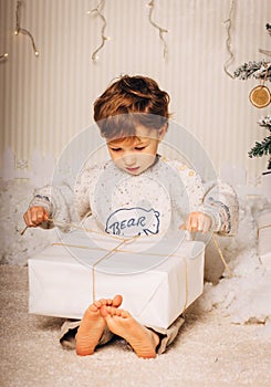 Cute little boy is sitting with a gift near the Christmas tree. The child is happy with New Year`s gift. Caucasian