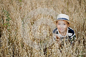 Cute little boy in a straw hat is sitting on an oat field playing hide and seek. Funny kid hiding in a field with oats.