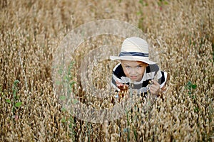 Cute little boy in a straw hat is sitting on an oat field playing hide and seek. Funny kid hiding in a field with oats.