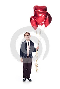 cute little boy in suit and eyeglasses holding red heart shaped balloons and smiling at camera