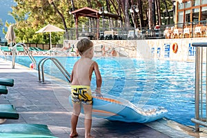 Cute little boy in swim trunks holds an inflatable swimming mattress at poolside in seaside resort