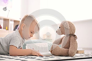 Cute little boy with teddy bear reading book on floor at home