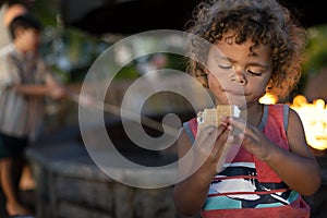 Cute little boy about to eat a s`more while sitting by an outdoor fire