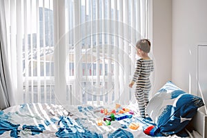 Cute little boy toddler standing on bed in room and looking into window waiting expecting someone. Child staring watching from