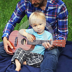 Cute little boy with ukulele is sitting on the lap of his father. Father teaches his son to play on the ukulele Hawaiian guitar