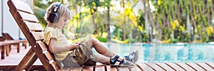 A cute little boy is using a smartphone and headphones lying on a deckchair by the pool. primary education, friendship, childhood,