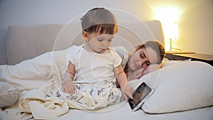 Cute little boy using tablet computer while mother is sleeping in bed