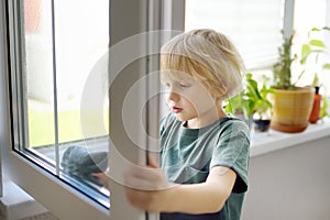 Cute little boy washing a window at home. Child helping parents with household chores, for example, cleaning windows in his house