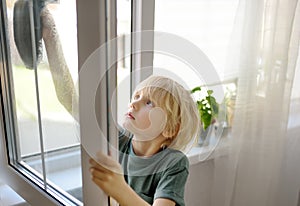 Cute little boy washing a window at home. Child helping parents with household chores, for example, cleaning windows in his house