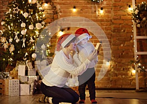 Cute little boy wearing Santa hat and his mother or grandmother regard a Christmas gift. Portrait of happy family on Christmas eve