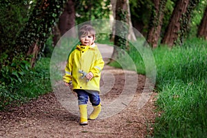 Cute little boy in yellow raincoat and rubber boots holding rhino toy with scared face walking alone lost in dark green forest.