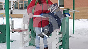 A cute little boy and a young mother are engaged in outdoor exercise equipment. It`s hard for a kid to reach the handles