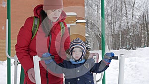 A cute little boy and a young mother are engaged in outdoor exercise equipment. It`s hard for a kid to reach the handles