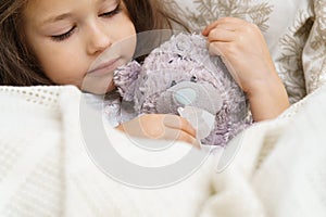 Cute little brunette girl healing, wiping imaginary snot of bear toy patient with tissues, lying, wrapped in blanket