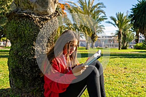 Cute little brunette girl reading a book outside on the grass. She is sitting at the base of a tree immersed in the