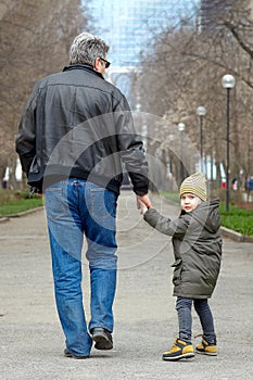 Cute little caucasial boy with blue eyes looks back walking with old-aged man, holding him with hand. Stroll in autumn park, casua