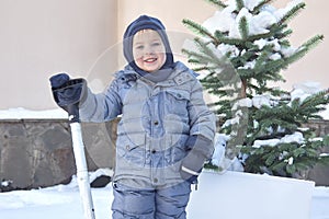 Cute little caucasian baby boy shovels snow in the yard with fir-tree on background. Winter outdoors, smiling, pink cheeks. Childr