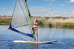 Cute little caucasian blond child girl enjoy having fun learning to surf on winsurfing board at freshwater pond lake or